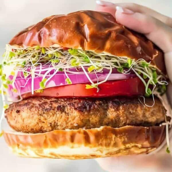 A sandwich with a grilled patty, sliced tomato, red onion, and sprouts in a toasted bun held in a hand.
