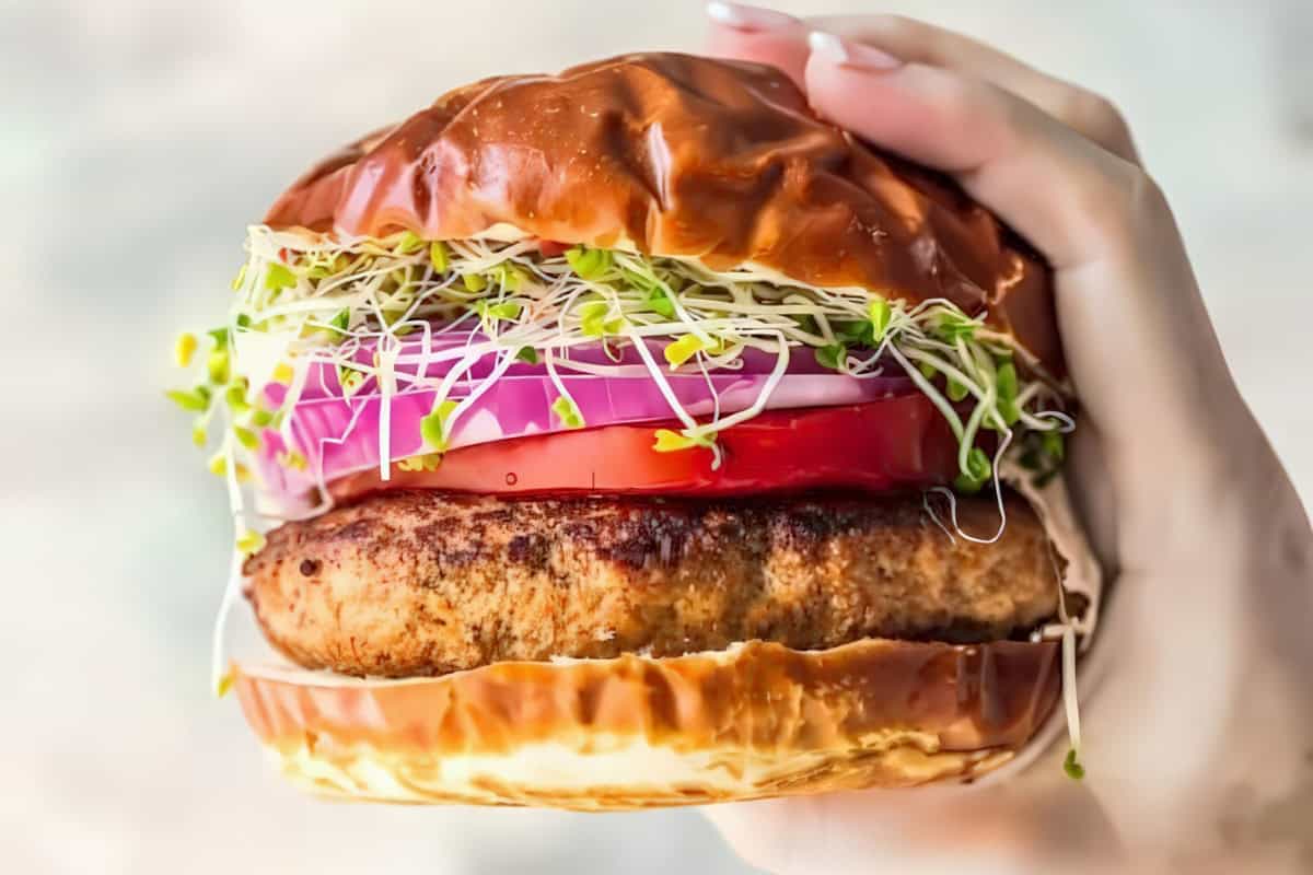 Close-up of a hand holding a well-made burger. The burger features a glossy, toasted bun and layers of fresh ingredients, including red onion rings, tomato slices, and a generous amount of sprouts, surrounding a juicy grilled patty.