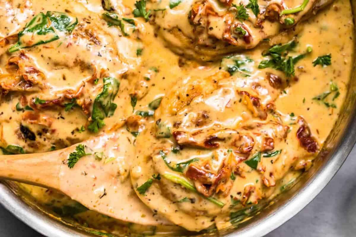 A close-up view of creamy Tuscan chicken in a skillet. The dish features succulent chicken breasts covered in a rich, golden sauce with sun-dried tomatoes, spinach, and freshly chopped parsley, all blended together to create a flavorful and visually appealing meal.