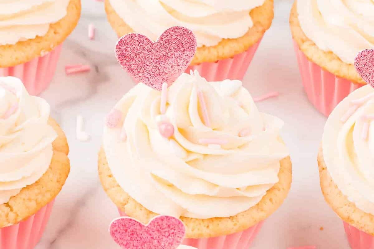 A cupcake with white frosting adorned with pink heart-shaped sprinkles and topped with a glittery pink heart. Other similarly decorated cupcakes are visible in the background.