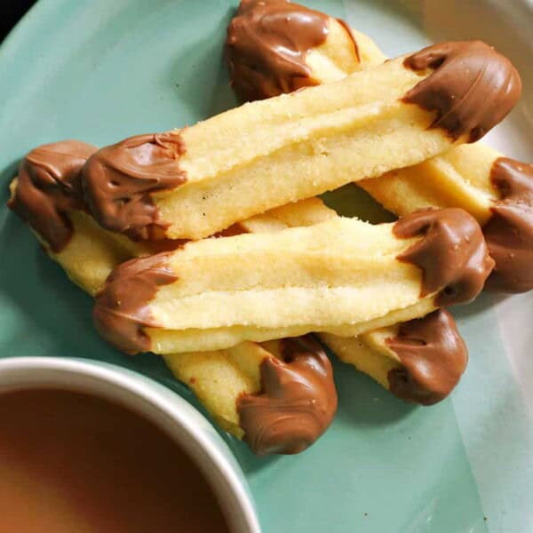 A plate of butter cookies with chocolate-dipped ends is next to a steaming cup of tea on a checkered cloth.