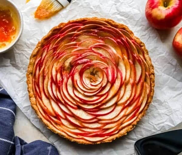 A freshly baked apple tart with thinly sliced apples arranged in a spiral pattern is placed on crumpled parchment paper. The tart is surrounded by a small bowl of apricot glaze, a brush, two whole apples, and part of a blue and white checkered cloth.
