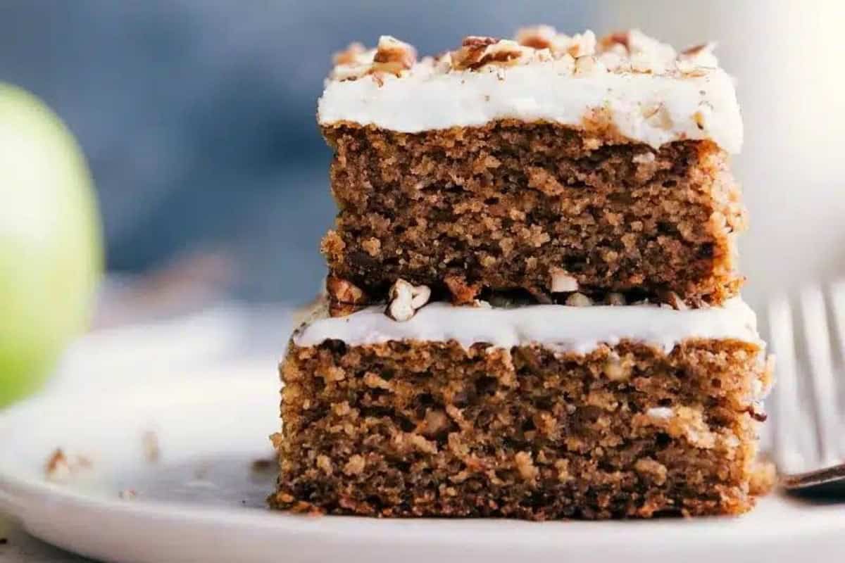Close-up of two stacked slices of cake topped with a layer of frosting and chopped pecans. The moist cake has a dark, slightly crumbly texture and is served on a white plate. A green apple and a fork are partially visible in the blurred background.