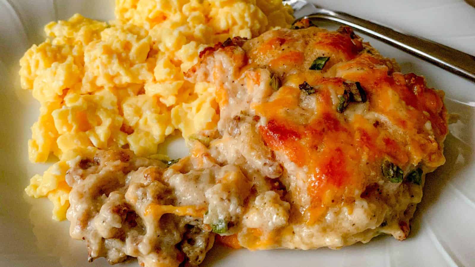 A plate of food featuring a serving of scrambled eggs and a portion of cheesy sausage casserole with green onion garnishes. A fork is placed next to the food on the right side of the plate.