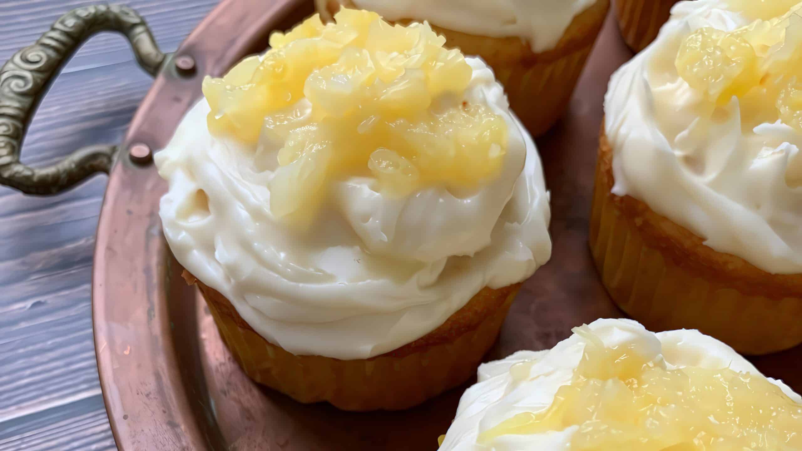 A close-up of several cupcakes topped with creamy white icing and a dollop of pineapple chunks. The cupcakes are placed on a copper tray with ornate handles, set against a wooden surface.