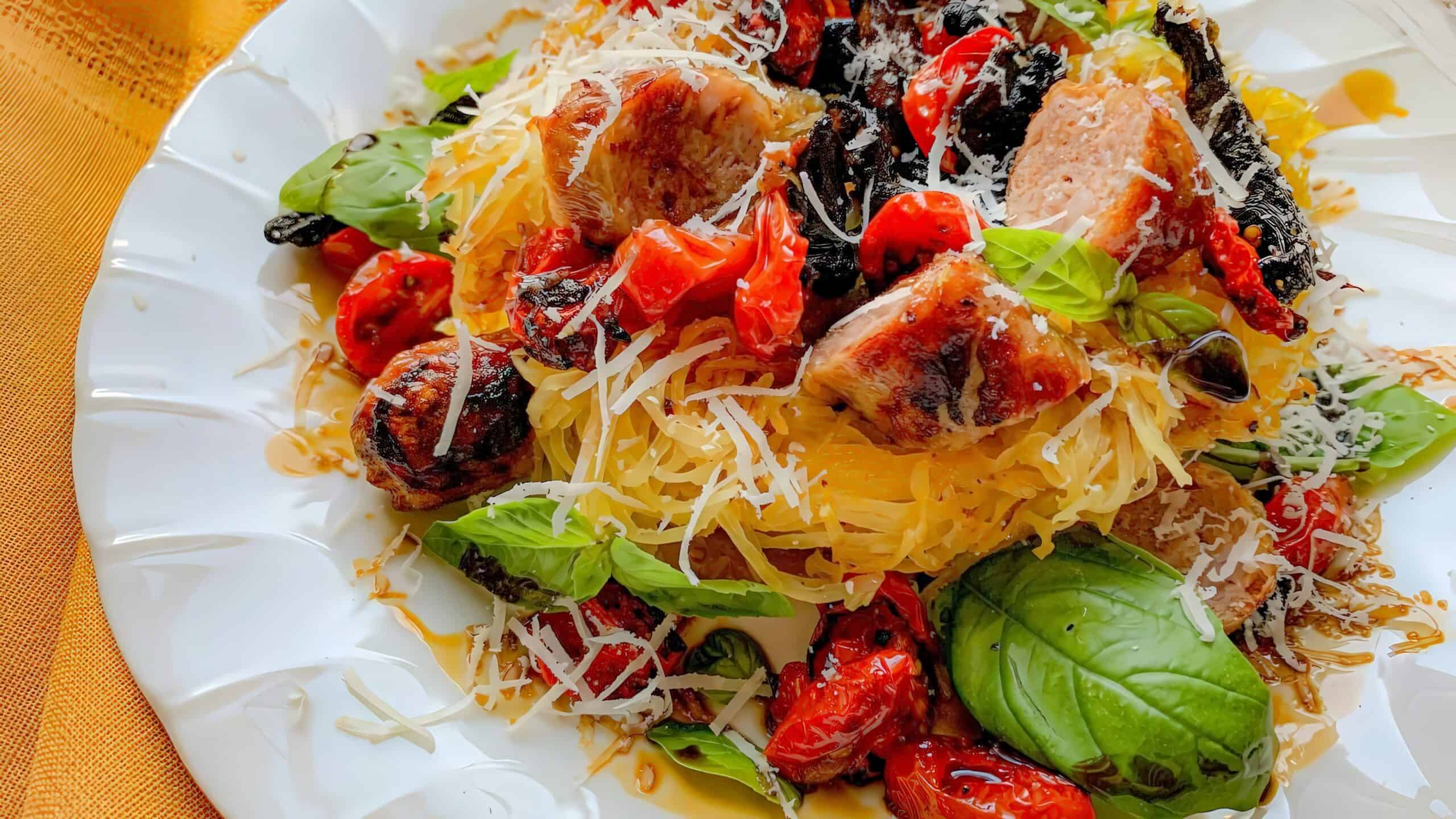 A plate of spaghetti squash topped with grilled sausage, cherry tomatoes, basil leaves, and shredded cheese.
