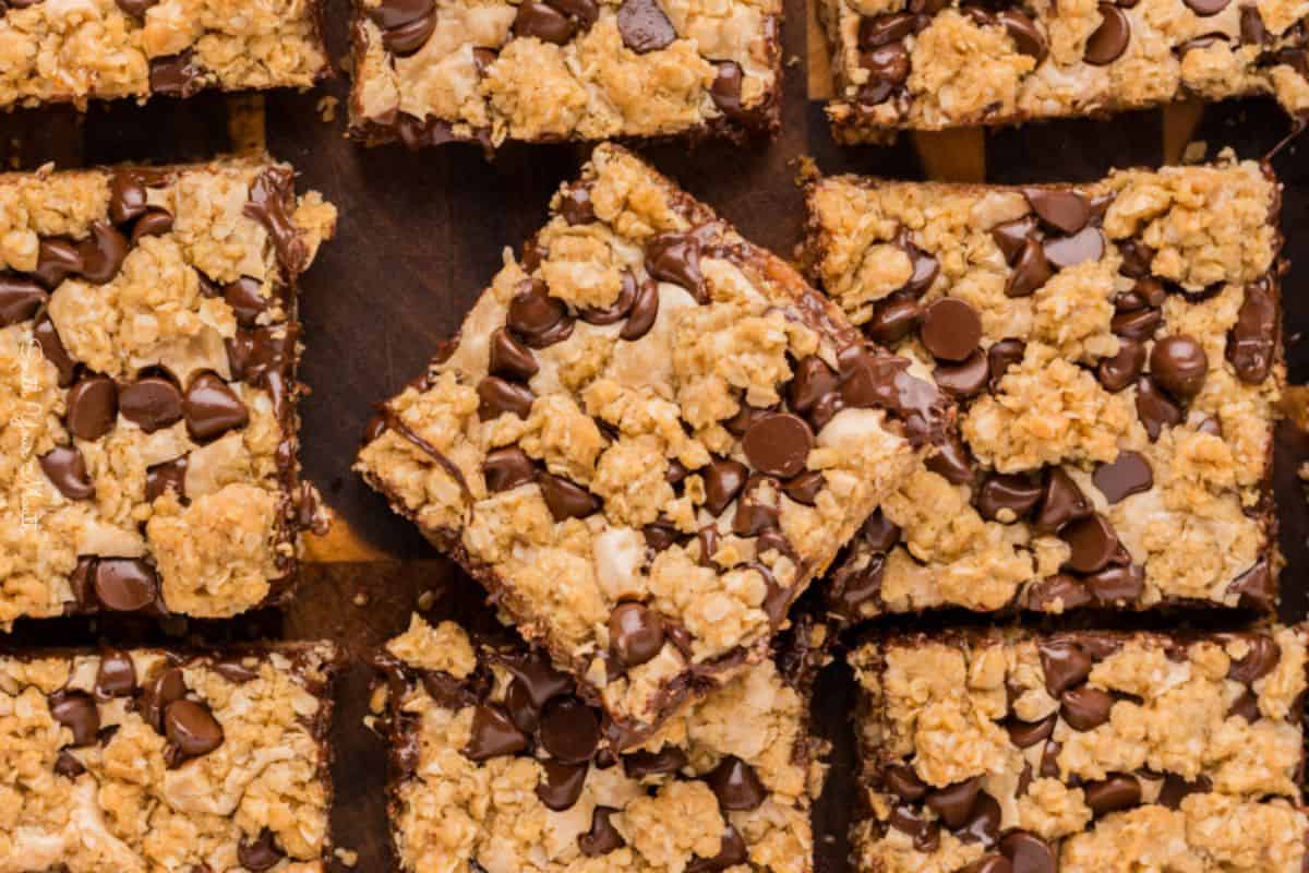 A close-up of several oat bars, topped with chocolate chips, arranged on a wooden surface. The bars have a crumbly texture with visible oats and melted chocolate, creating a tempting, homemade dessert appearance.