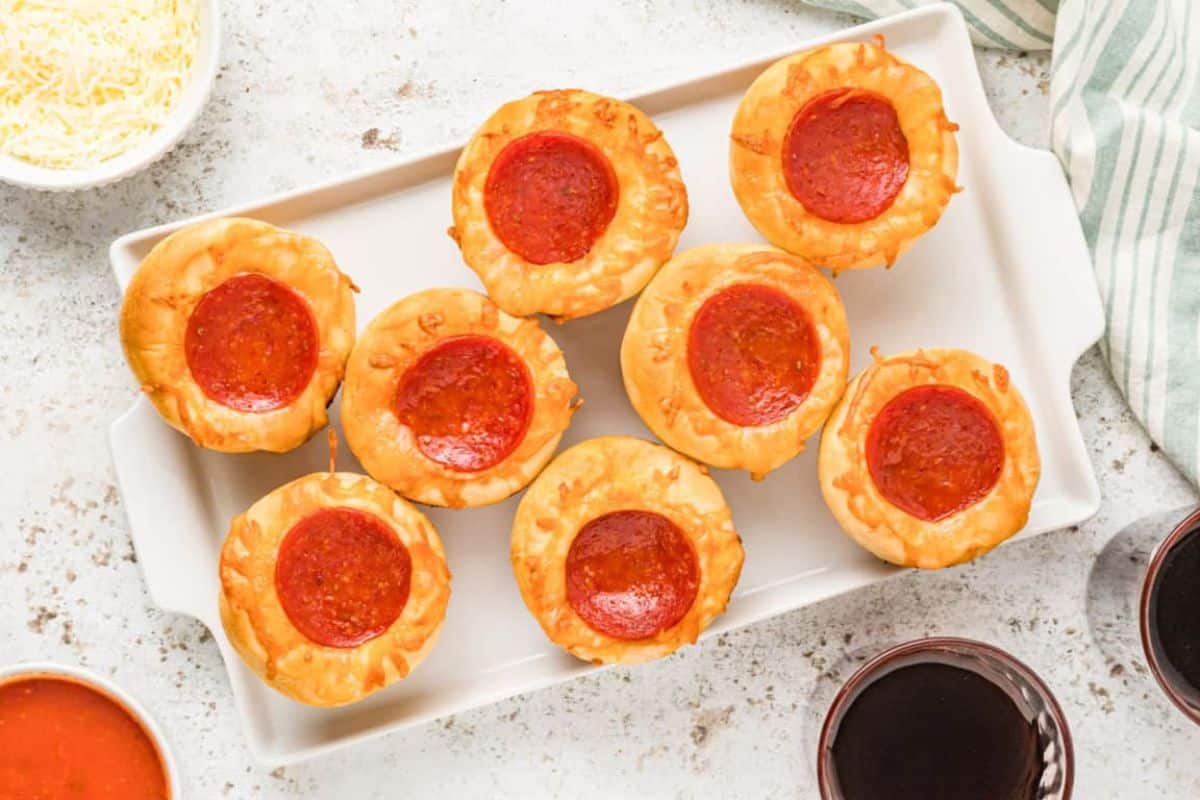 A white rectangular plate holds eight mini pepperoni pizza cups, each topped with a slice of pepperoni and melted cheese. Surrounding the plate are bowls with shredded cheese and pizza sauce, and two glasses of dark beverage. A striped napkin is placed on the side.