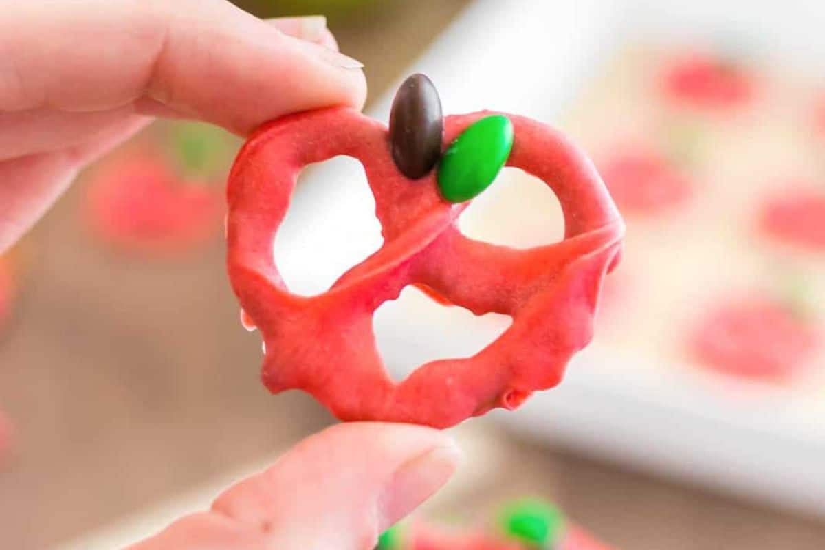 A close-up of a hand holding a pretzel dipped in red candy coating, shaped to resemble an apple. The pretzel is topped with a green candy resembling a leaf and a brown candy resembling a stem. Blurry pretzels on a tray are visible in the background.