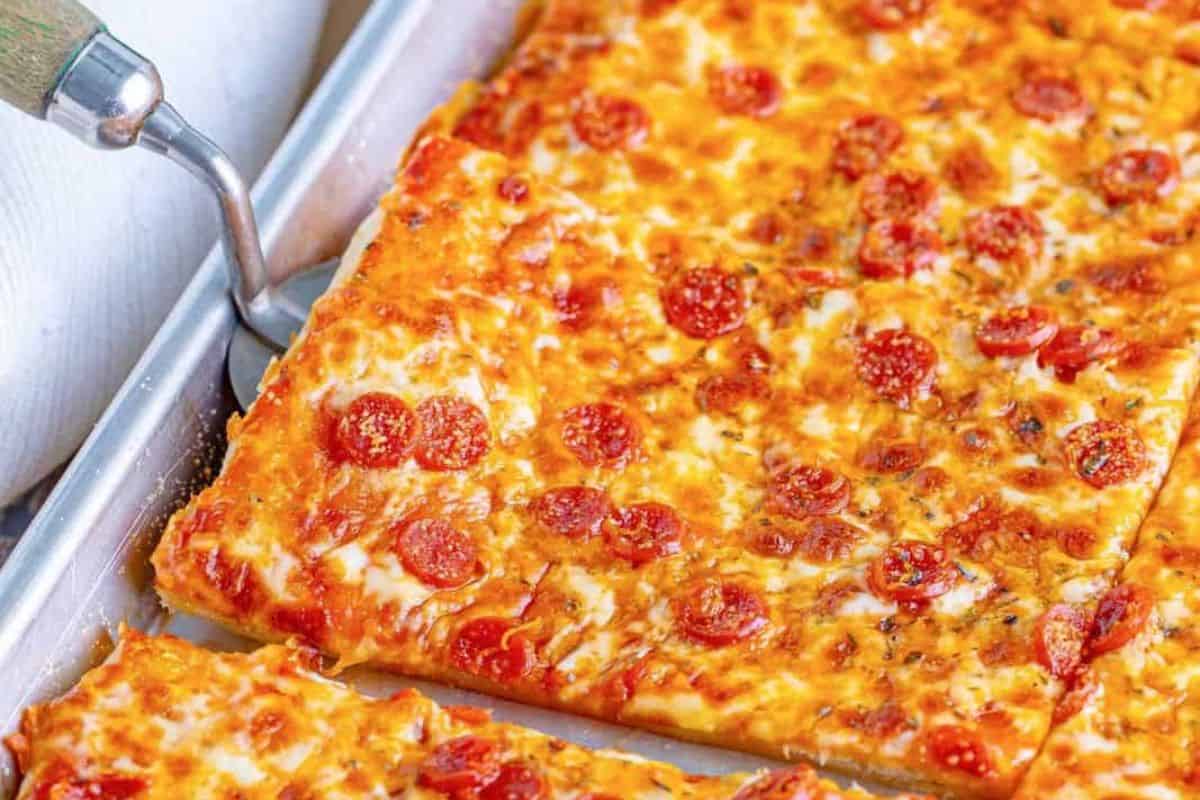 A tray of freshly baked pepperoni pizza cut into rectangular slices. The pizza has a golden-brown crust, melted cheese, and pepperoni pieces evenly distributed on top. A pizza cutter is partially visible, lifting one of the slices.