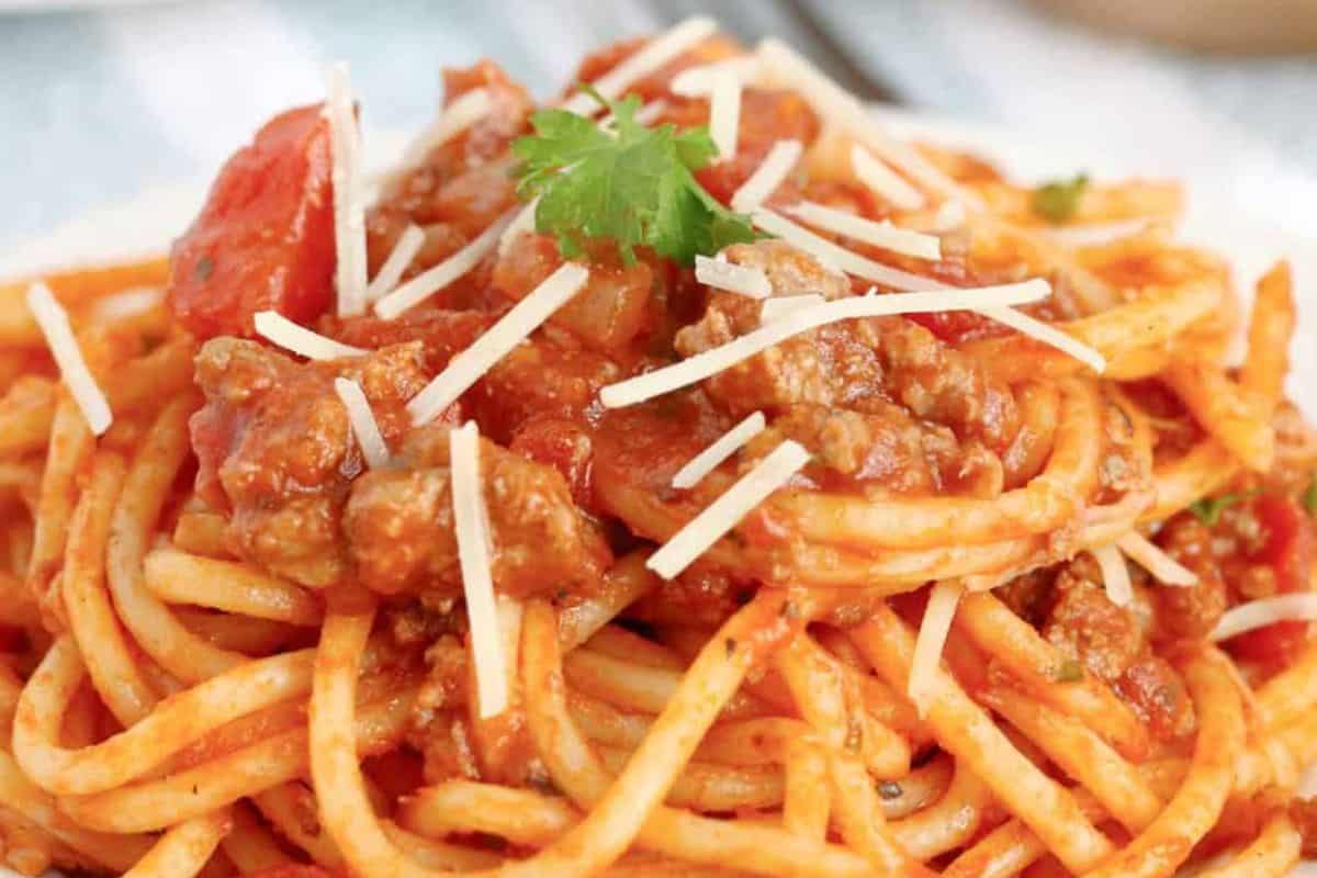 A close-up of a plate of spaghetti bolognese. The pasta is topped with a rich meat sauce, grated parmesan cheese, and garnished with a sprig of parsley.