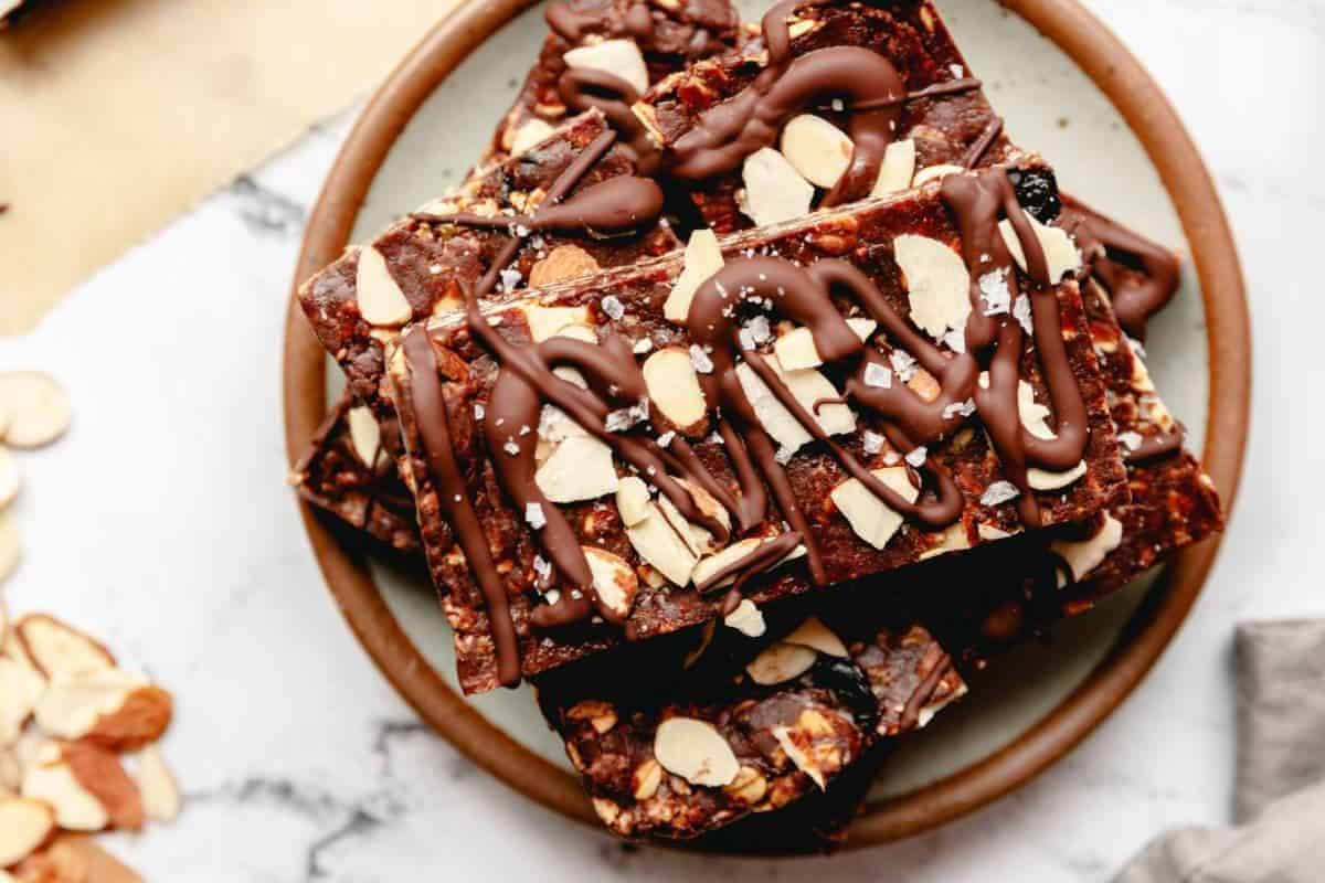 A plate of chocolate almond bark squares topped with sliced almonds and drizzled with melted chocolate, viewed from above. The bark is rich and dark, with a slightly rough texture and a few grains of sea salt scattered on top.