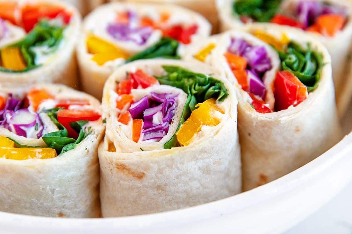 Close-up of a platter of fresh veggie wraps, cut into pieces. Each wrap includes colorful layers of red, orange, and yellow bell peppers, purple cabbage, and leafy greens, all wrapped in soft tortilla bread. The vibrant colors create a visually appealing presentation.