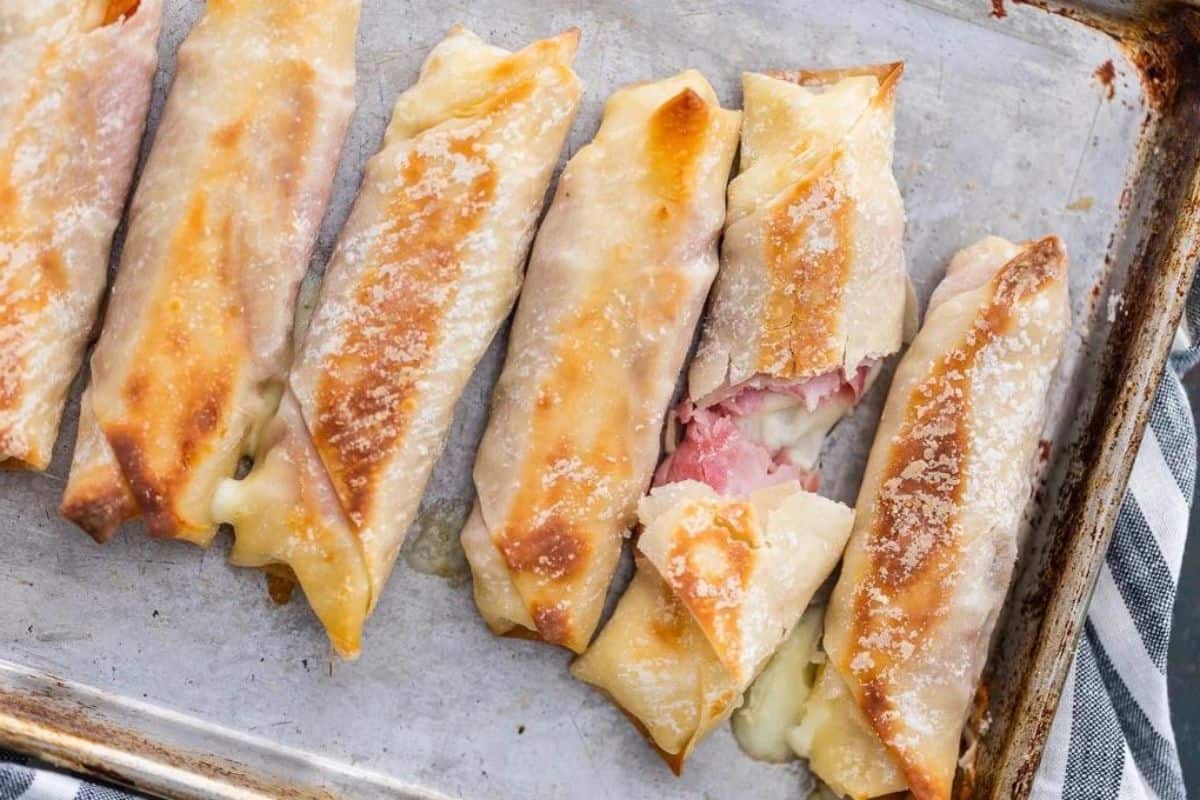 A baking tray holds several baked egg rolls with golden, crispy exteriors. One roll is slightly open, revealing a filling of melted cheese and ham. A striped cloth napkin is visible at the corner of the tray.