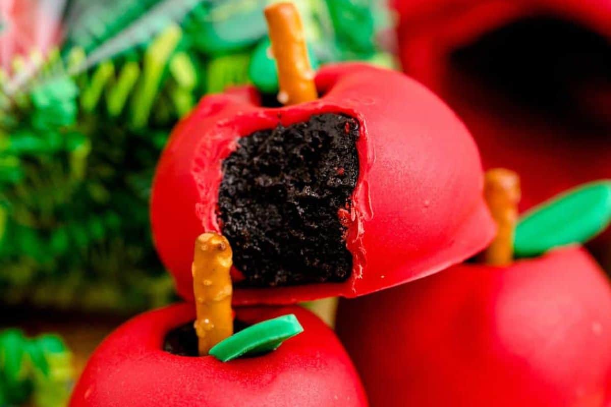 Close-up of two treats shaped like bright red apples with a green leaf and a pretzel stick as the stem. The apple in front has a bite taken out, revealing a dark chocolate filling inside. In the background, blurred greenery and another red item are visible.