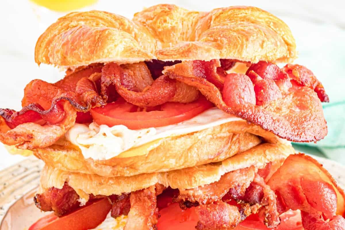 A close-up of a croissant sandwich filled with crispy bacon strips, fresh tomato slices, and a fried egg. The sandwich is layered, highlighting its hearty and savory ingredients, with a golden-brown croissant as the base.
