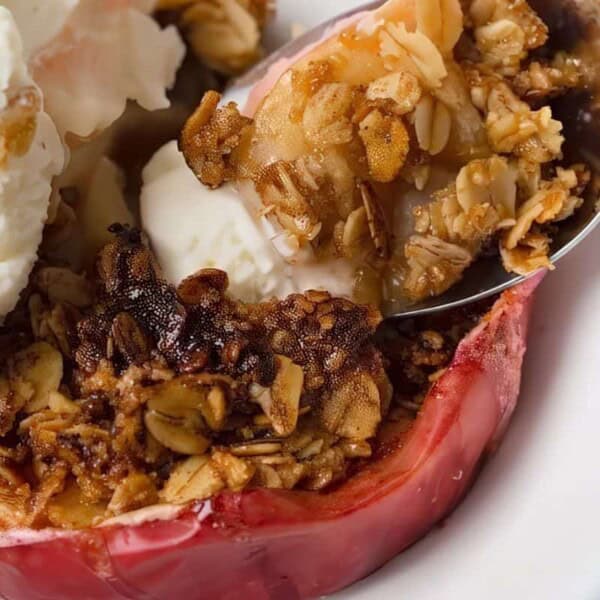 A close-up of a spoonful of baked apple crisp with oats and a scoop of vanilla ice cream on the side on a white plate.