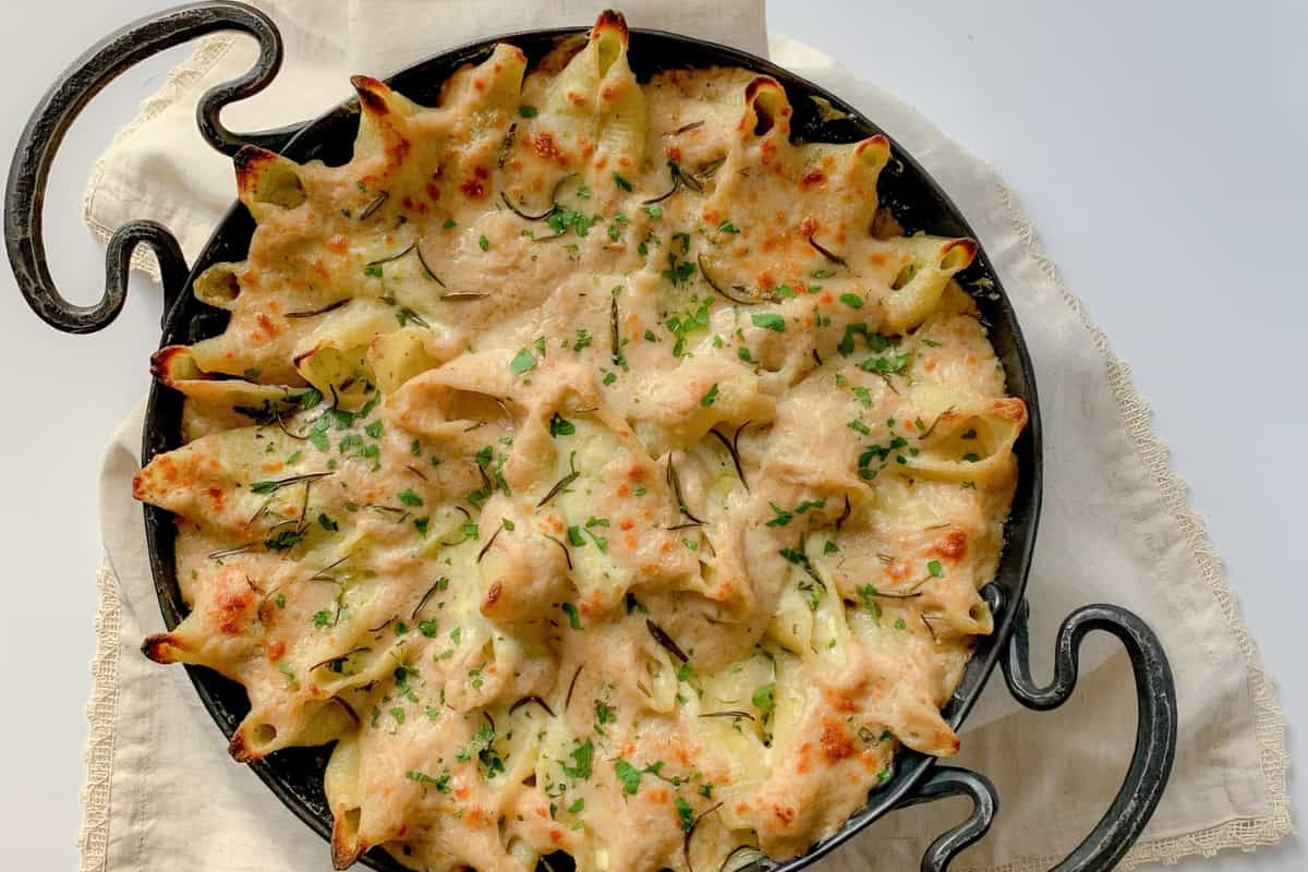 A round baking dish filled with baked pasta shells topped with melted cheese and garnished with chopped herbs. The dish sits on a cream-colored cloth, and the pasta appears golden brown and crispy on top.