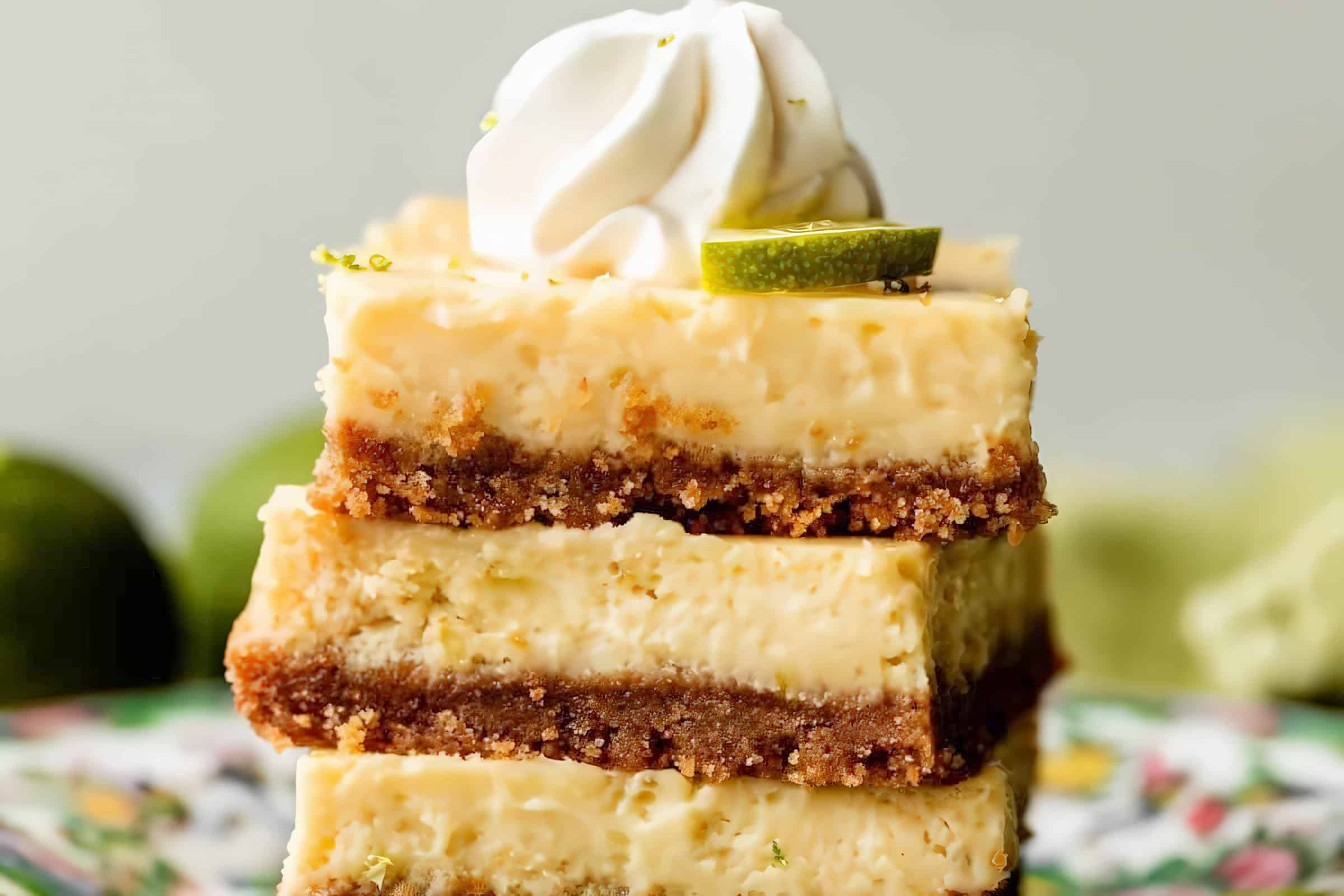 A stack of three key lime pie bars with graham cracker crust, garnished with whipped cream and a slice of lime on top. The bars have a creamy yellow filling and are set against a blurred background with green and colorful elements.