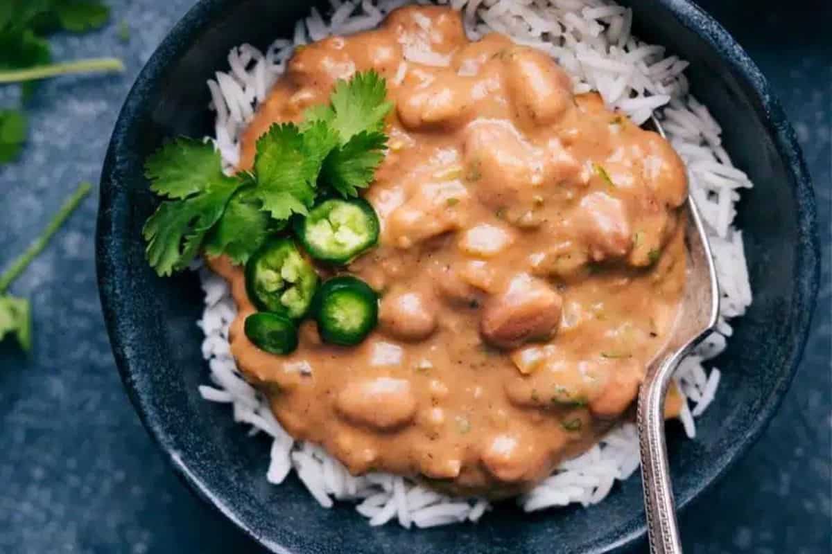 A bowl of white rice topped with a creamy bean curry garnished with cilantro and sliced green chilies. A spoon is placed in the bowl. The dish is presented on a dark surface with loose cilantro leaves nearby.