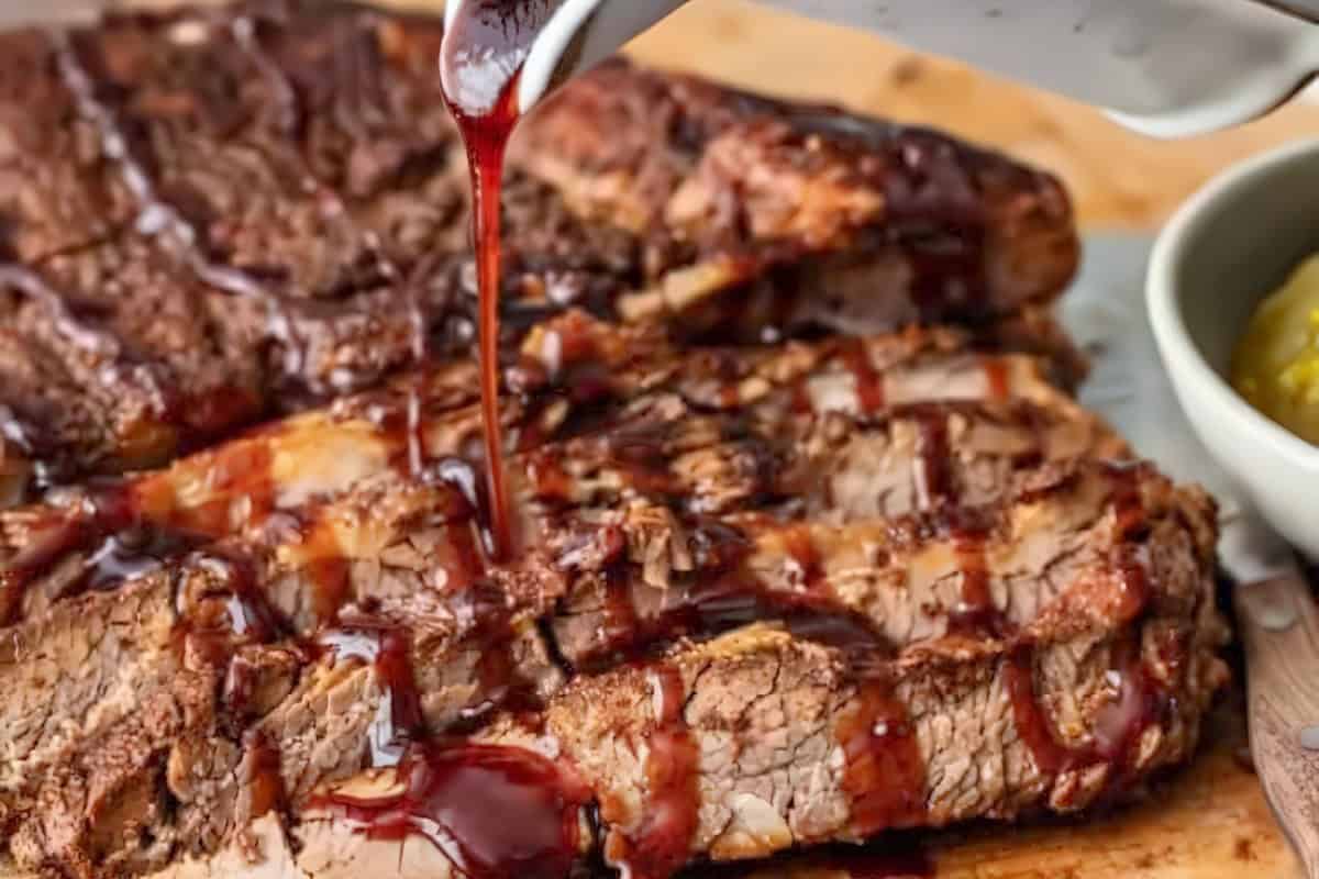 Close-up of grilled meat slices on a wooden surface being drizzled with barbecue sauce from a metallic container. The dish appears well-seasoned and cooked, with a bowl of pickles visible in the background.