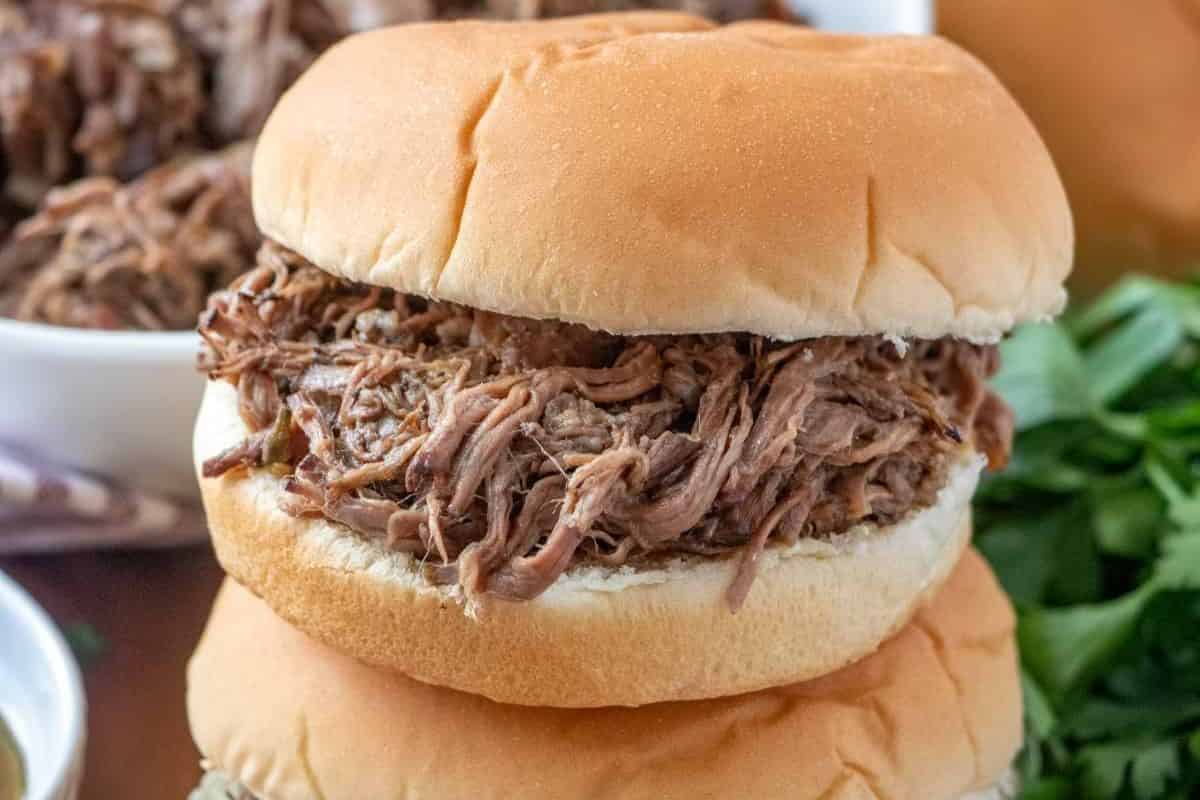 A close-up of a pulled pork sandwich with shredded pork overflowing from a soft, golden-brown bun. The background shows a bowl of more pulled pork and some greens, adding to the fresh and hearty appearance of the meal.