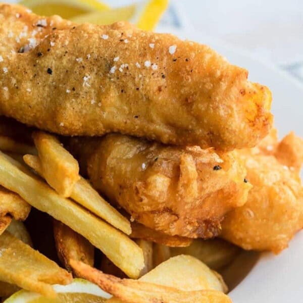 Close-up of a plate of fish and chips. The dish features two pieces of golden-brown, battered and fried fish fillets placed on a bed of thick-cut French fries. The fish is sprinkled with coarse salt and pepper. A few lemon wedges are visible in the background.