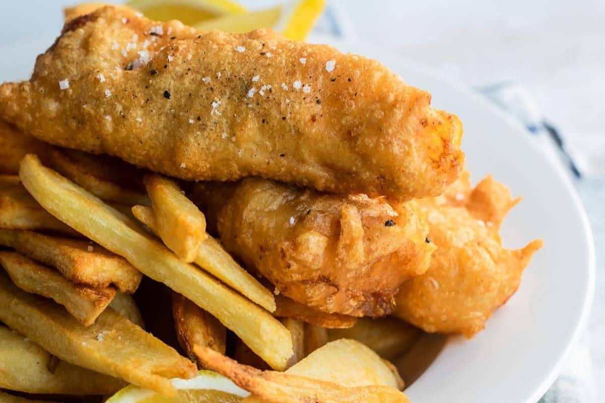 Close-up of a plate of fish and chips. The dish features two pieces of golden-brown, battered and fried fish fillets placed on a bed of thick-cut French fries. The fish is sprinkled with coarse salt and pepper. A few lemon wedges are visible in the background.