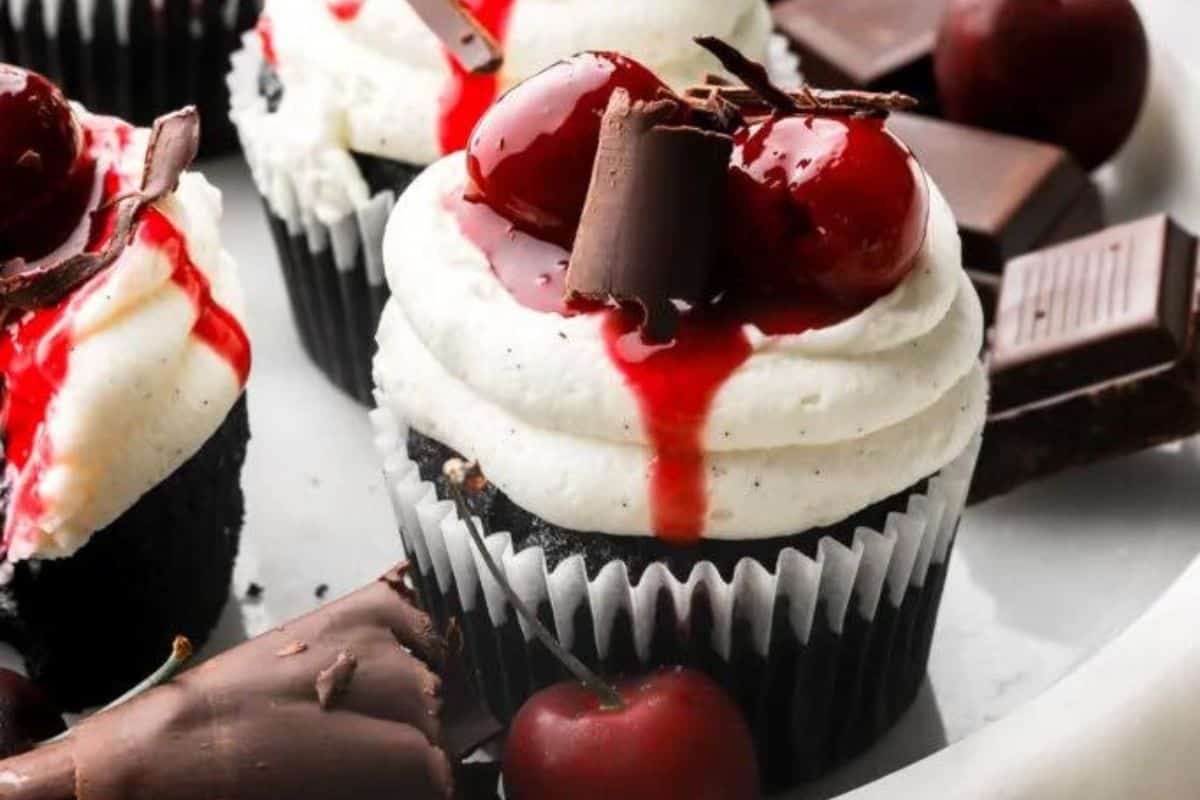 A close-up of a chocolate cupcake topped with swirls of white frosting, juicy red cherries, chocolate shavings, and a drizzle of red sauce. Pieces of dark chocolate and fresh cherries are placed around the cupcake on a white plate.