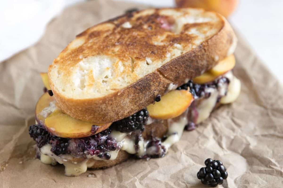 A grilled sandwich with melted cheese, sliced peaches, and blackberries is placed on brown parchment paper, with a fresh blackberry in the foreground. The bread appears golden and crispy from grilling.