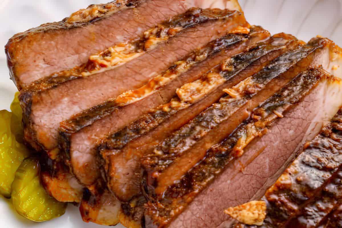 Close-up of thick slices of smoked brisket with a well-seasoned crust, arranged on a plate with a few pickle slices on the side. The meat appears tender and juicy, with visible marbling and layers of seasoning.