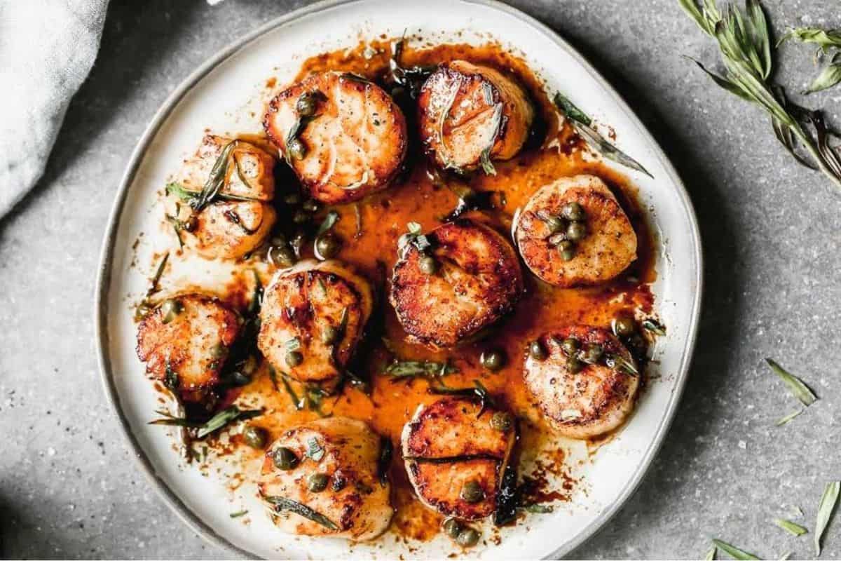 A plate of seared scallops garnished with capers and fresh herbs, drizzled with a brown butter sauce. The scallops are golden brown and attractively arranged on a light-colored plate set on a gray surface. A sprig of herbs is partially visible in the background.