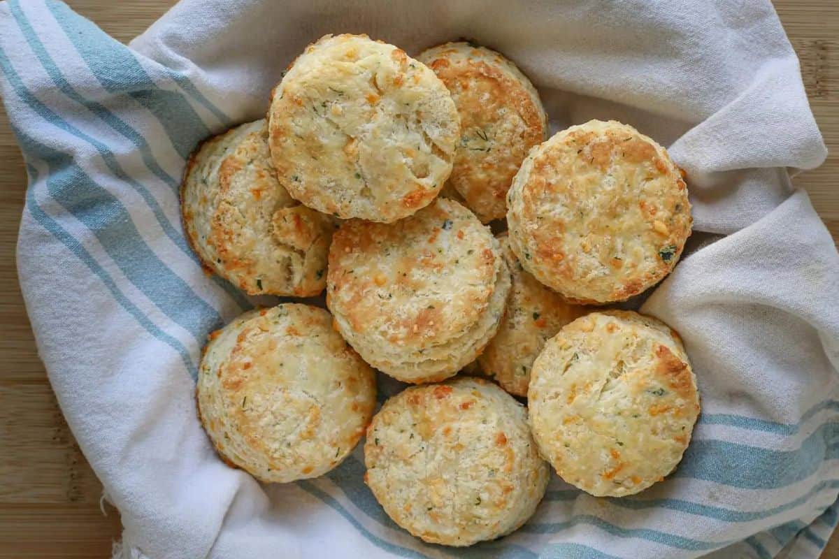 A cloth-lined basket filled with freshly baked cheddar cheese biscuits, golden and slightly browned on top, sitting on a wooden surface.