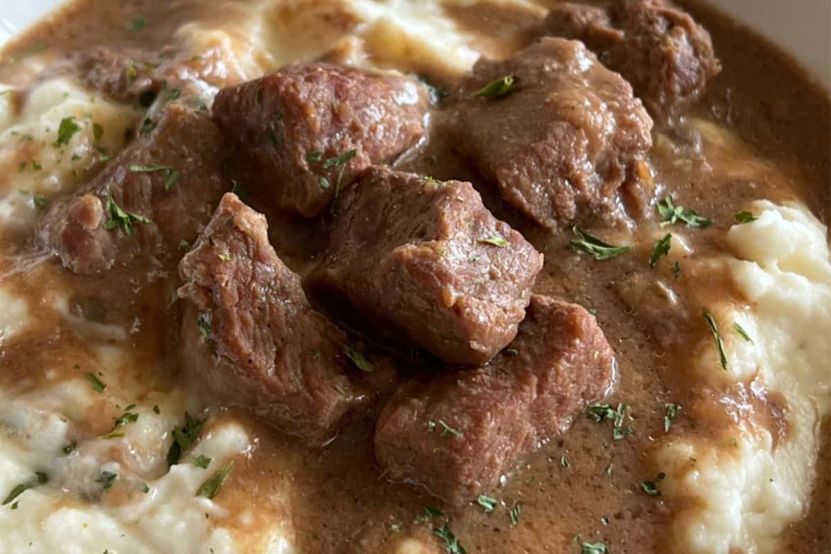 A close-up shot of a bowl of beef stew served over creamy mashed potatoes. The stew features tender chunks of beef in a rich brown gravy, garnished with chopped herbs. The mashed potatoes appear smooth and fluffy, complementing the hearty stew.