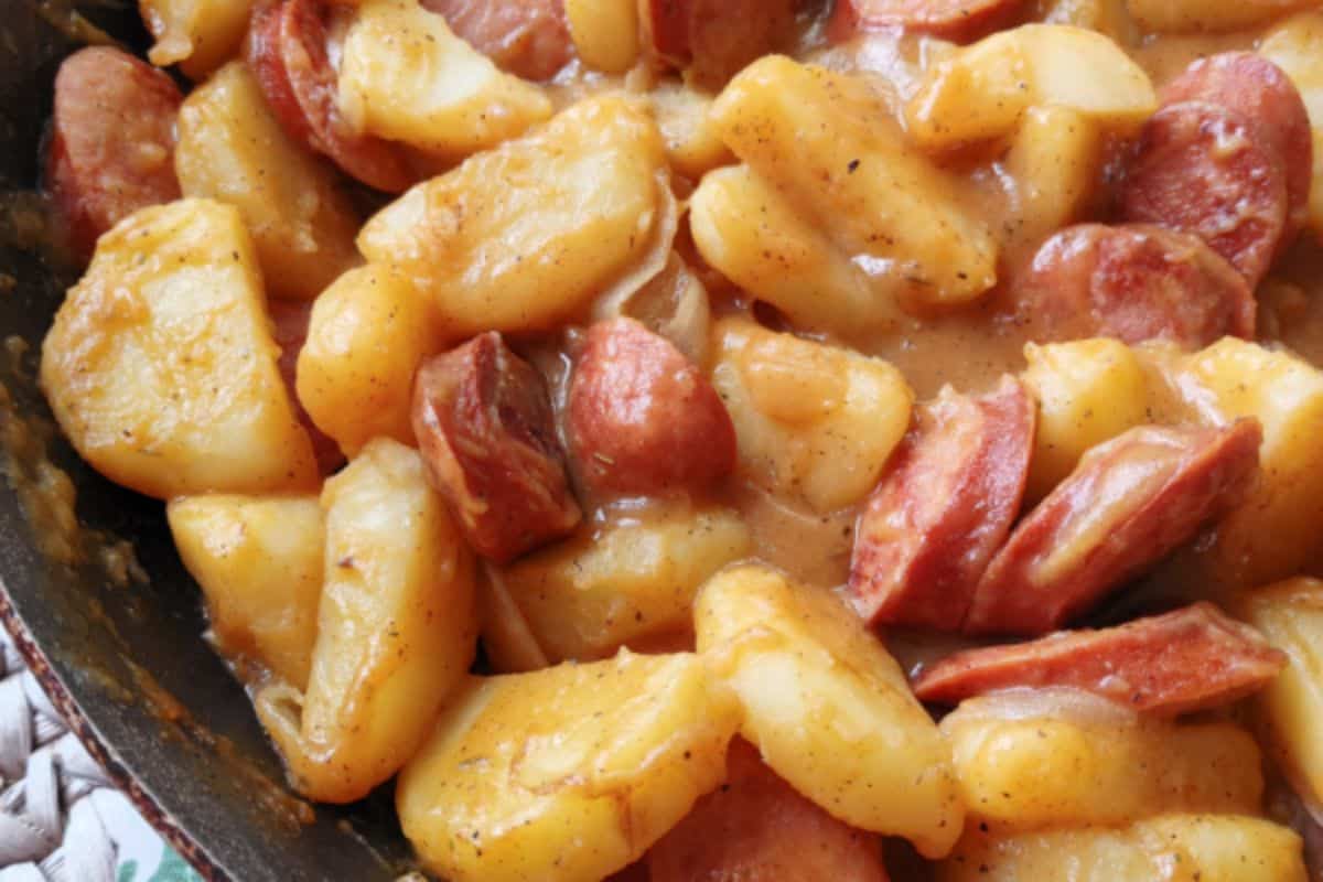 A close-up of a hearty dish composed of cooked sliced potatoes and chunks of sausage, all coated in a savory sauce. The dish appears to be rich and flavorful, with a golden-brown hue.