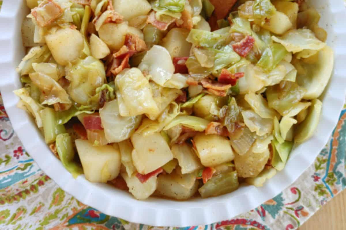 A close-up of a white bowl filled with a savory dish of cooked cabbage, diced potatoes, and bacon pieces. The bowl is placed on a colorful, patterned tablecloth, adding a vibrant touch to the overall presentation of the food.