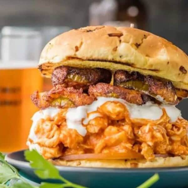 A fried chicken sandwich topped with plantains and creamy sauce, served on a toasted bun. A glass of beer sits in the background.
