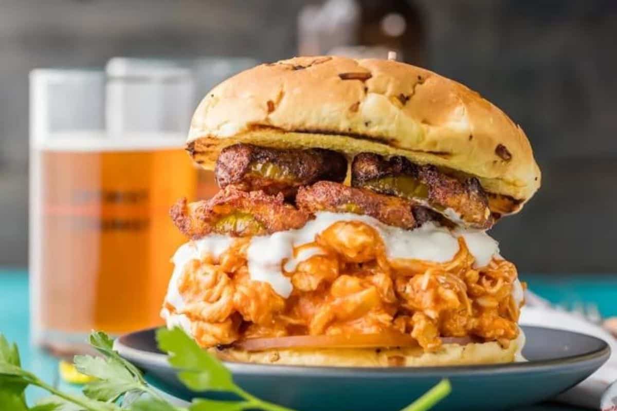 A fried chicken sandwich topped with plantains and creamy sauce, served on a toasted bun. A glass of beer sits in the background.