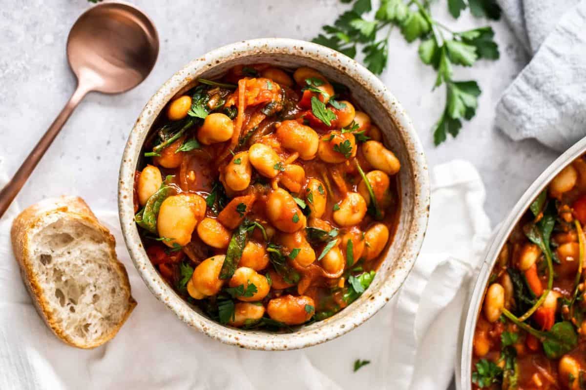 A bowl of hearty bean stew, featuring white beans, spinach, and a rich tomato sauce, garnished with fresh parsley. Next to the bowl is a piece of crusty bread on a table, along with a rustic bronze spoon and sprigs of parsley.