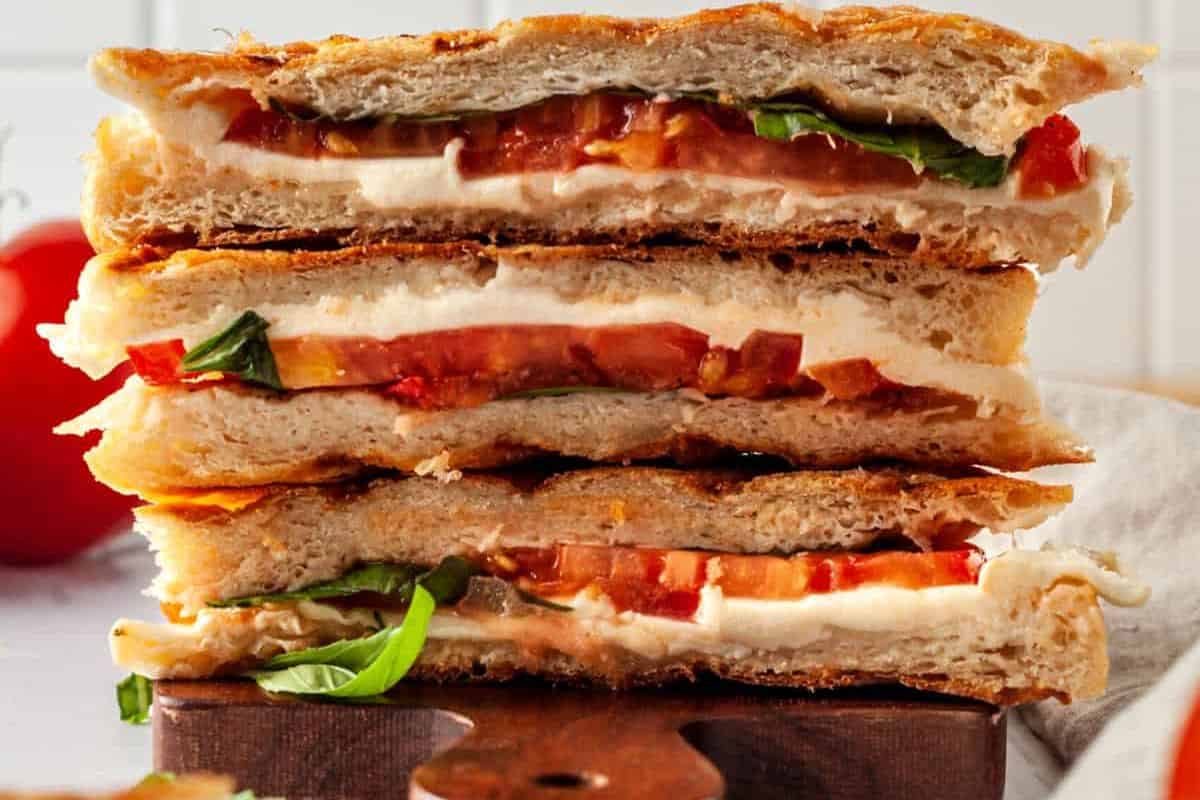A stack of three Caprese sandwiches filled with fresh mozzarella cheese, ripe tomato slices, and basil leaves, served on toasted ciabatta bread. The layers of the sandwich are clearly visible, showing the fresh ingredients. The sandwiches are placed on a wooden board.