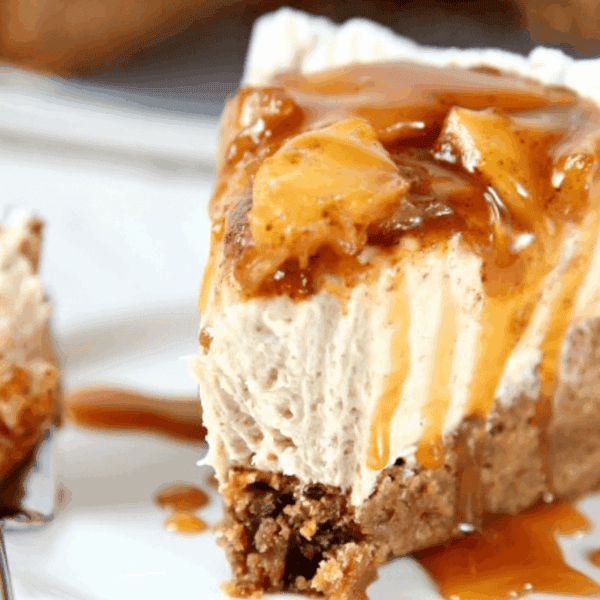 A slice of cheesecake topped with caramel sauce sits on a plate with fruit and a fork holding a small bite nearby.