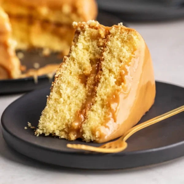 A slice of caramel cake with golden-brown layers and a smooth caramel frosting is placed on a black plate with a gold fork beside it. The remaining cake, partially visible in the background, is also frosted with caramel.