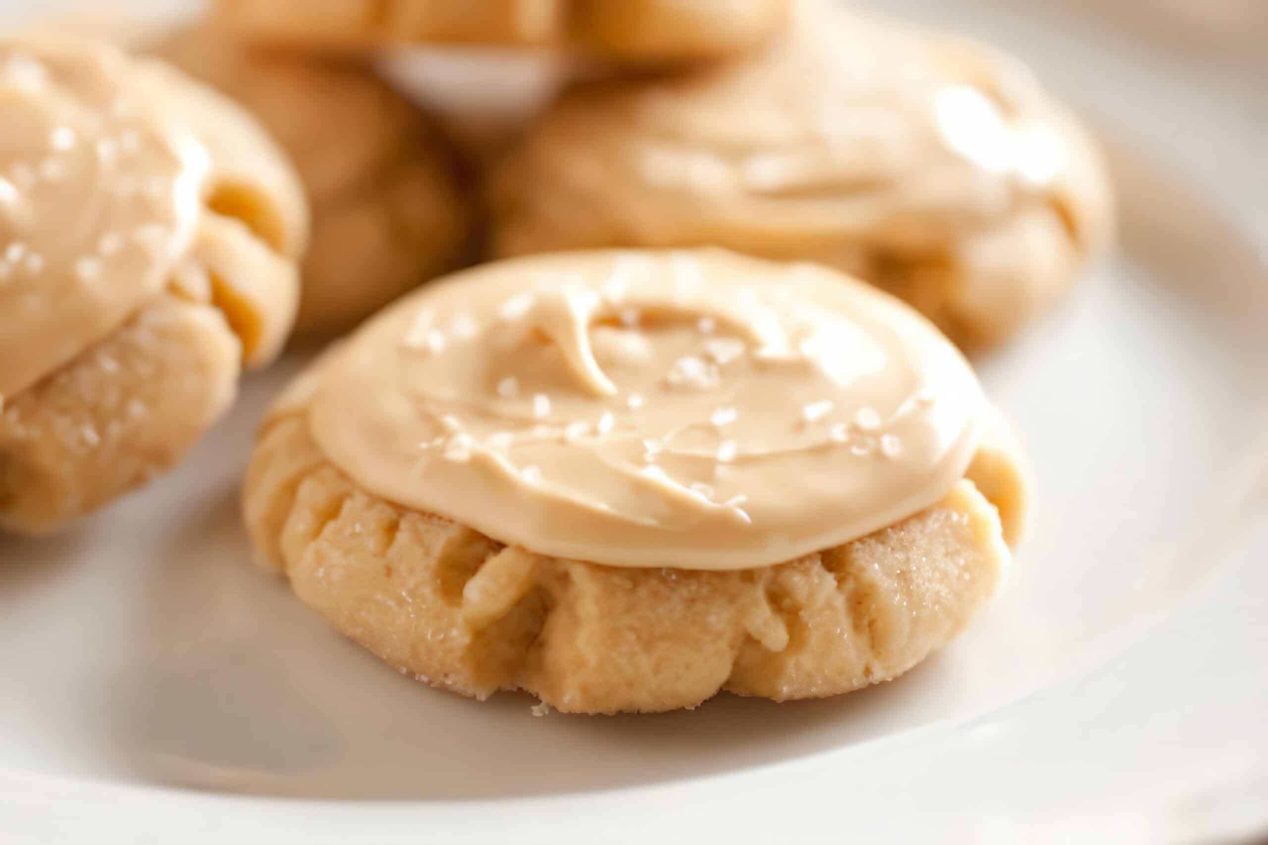 Close-up of a creamy frosted cookie on a white plate. The cookie has a round shape with a slightly textured edge and is topped with a smooth layer of caramel-colored frosting. Other similar cookies are blurred in the background.