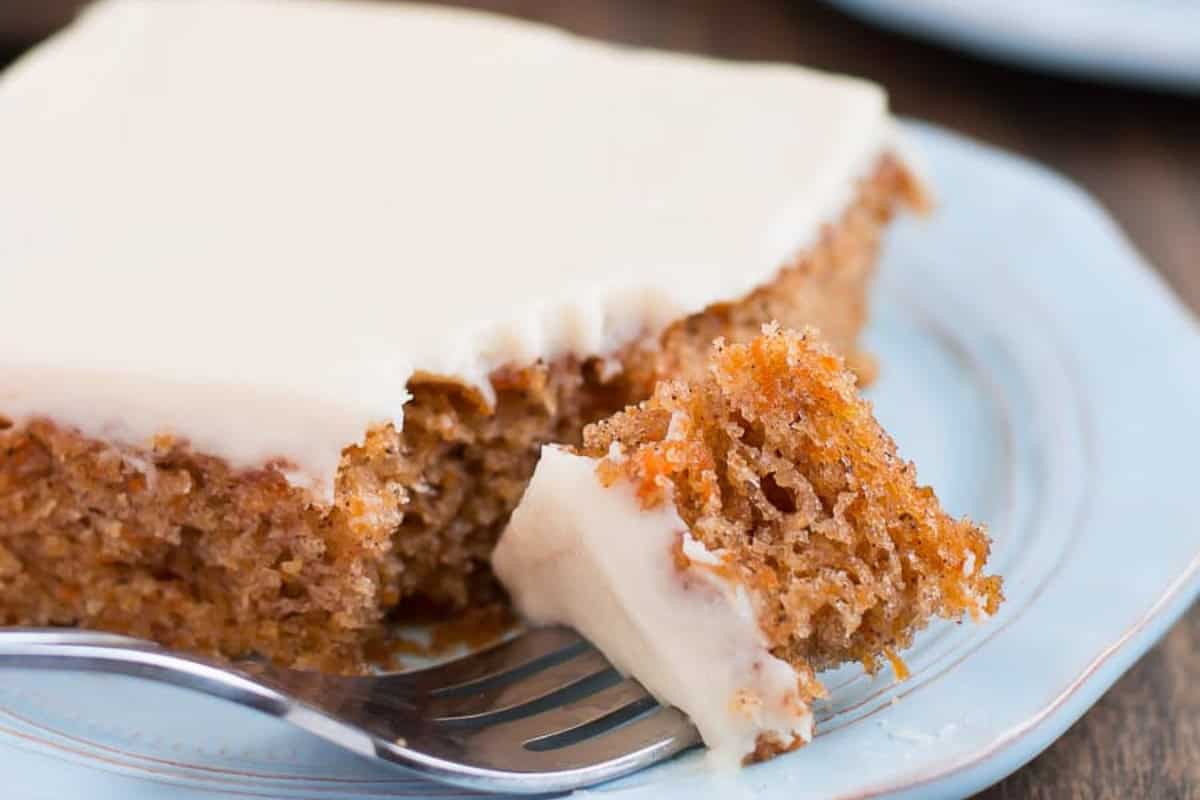 A close-up of a slice of carrot cake on a light blue plate. The cake has a thick layer of white cream cheese frosting on top. A fork is lifting a small piece from the slice, showing the moist, textured interior with bits of carrot visible.