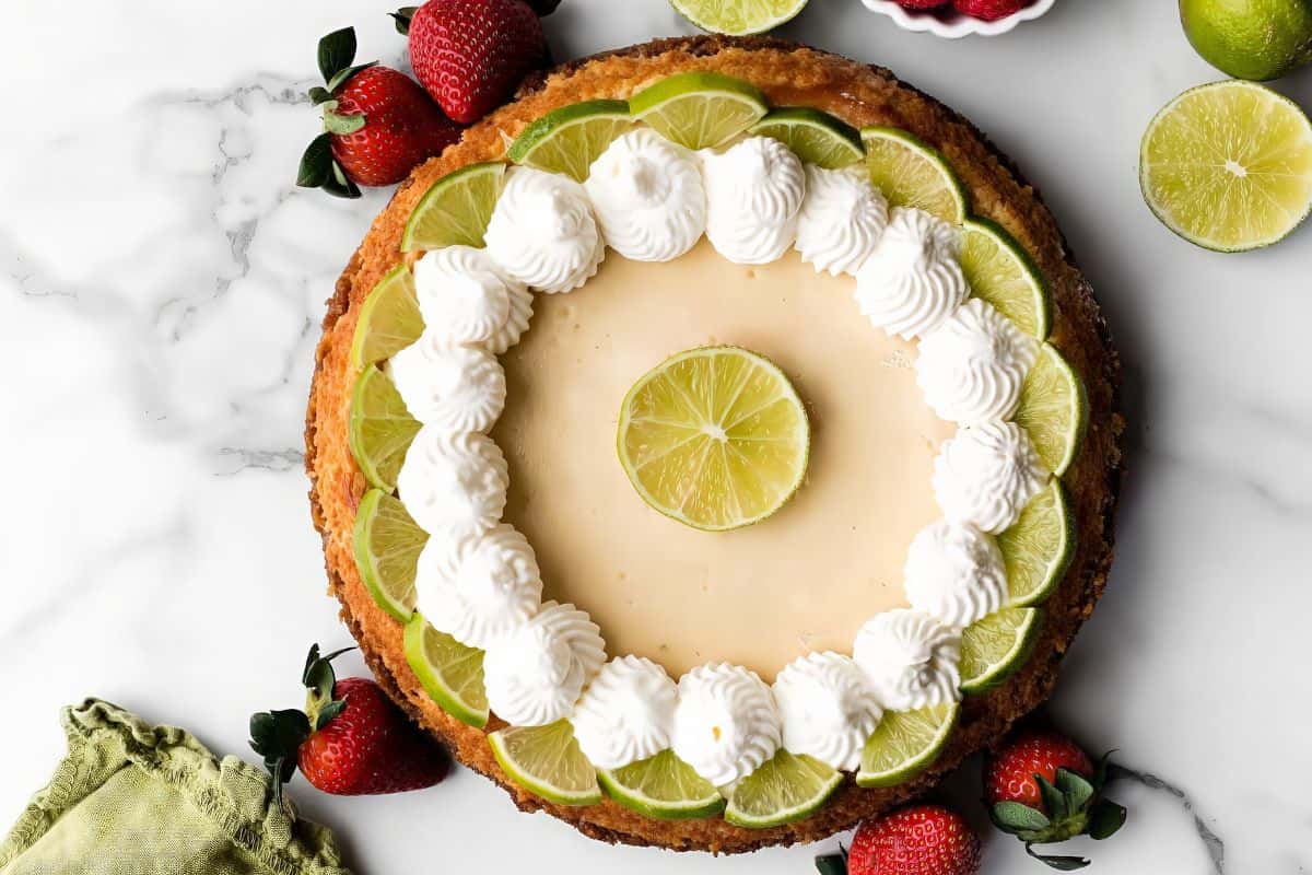 A top view of a key lime pie garnished with a ring of lime slices and dollops of whipped cream on top, with a lime slice in the center. Fresh strawberries and whole limes are scattered around the pie on a white marble surface. A green napkin is placed to the side.