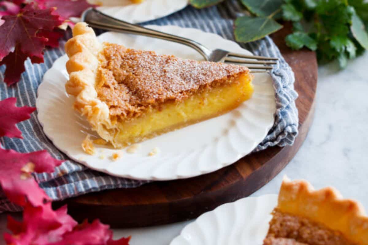 A slice of custard pie with a golden, flaky crust is served on a white plate, accompanied by a fork. The pie is garnished with a light dusting of powdered sugar, and is placed on a decorative cloth, surrounded by autumn leaves and greenery.