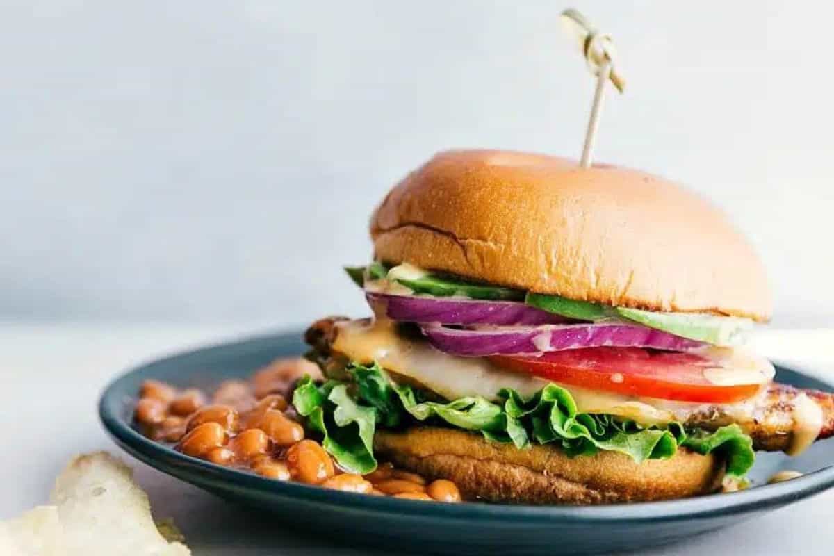 A grilled chicken sandwich with fresh lettuce, tomato, red onion, avocado, and melted cheese on a toasted bun. It is held together with a skewer and served on a black plate next to a side of baked beans.