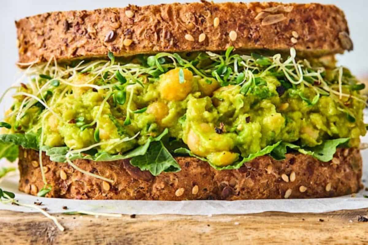 A close-up of a sandwich made with whole grain bread, filled with mashed avocado, chickpeas, leafy greens, and sprouts. The sandwich is placed on a piece of parchment paper on a wooden surface.