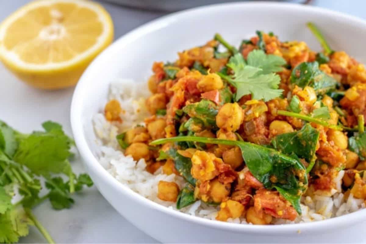 A white bowl filled with a vibrant chickpea and spinach curry served over white rice, garnished with fresh cilantro. A halved lemon and sprigs of cilantro are placed next to the bowl on a light-colored surface.