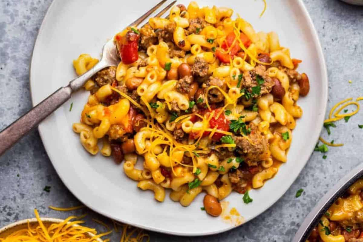 A plate of chili macaroni, topped with ground beef, beans, diced tomatoes, and shredded cheddar cheese. A fork is placed on the side of the plate, and some shredded cheddar cheese is scattered next to the plate on a gray surface.